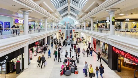 Metrocentre The inside of the Metrocentre. Shoppers walk around and sit on seats in the middle of the hall. Shops such as Superdrug and JD line the hallway.