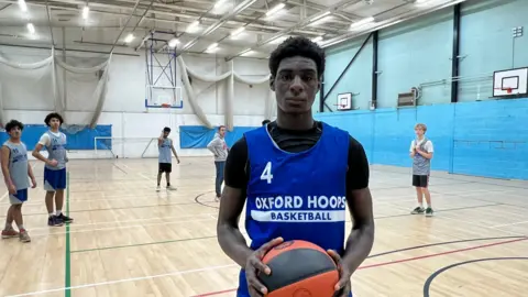 David Okerike faces the camera. He's wearing a blue vest with 'Oxford Hoops' printed on it in white letters. He's holding an orange and black basketball in both hands. Behind him is a school hall, with a wooden panelled floor marked for various sports and several basket ball hoops fixed to the walls. Behind David there are several other young players dotted around the court looking in the direction of the camera.