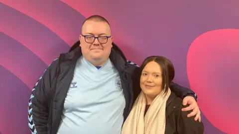Dave Shephard and Simone Seth :  A clean-shaven man with a shaved dark hair is wearing a blue tshirt and dark blue sports jacket with his arm around a shorter woman with pulled back dark hair and a beige scarf. They are standing against a swirly purple wall