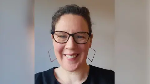 Lucy is looking at the camera and can be seen from the shoulders up. She's wearing large square-hooped earrings and smiling, with her hair back.