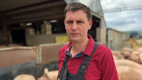 Danny Skinner, wearing a red sweatshirt and dark blue dungarees, in from of his pigs.