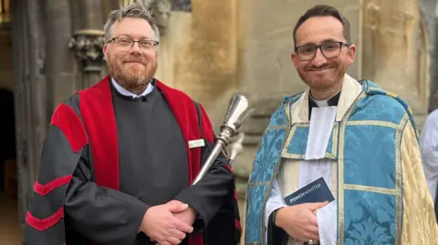 Jon Wright/BBC Jim Robinson, verger at Ipswich Minster, and Revd Tom Mumford, both dressed in Anglican church robes, stand outside St Mary Le Tower church