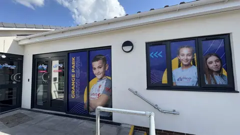 The entrance to Grange Park Youth Centre, a single storey, white rendered building with large black-framed windows and blue doors that have full length photos of smiling young people.
