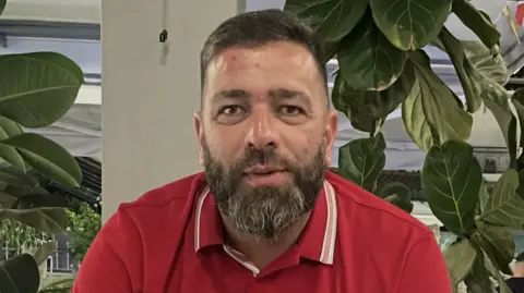 Gary Porritt is wearing a red polo style t-shirt and has a brown bears with white streaks. He has short brown hair and is sitting in front of a white pillar and green plants. 