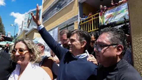 EPA/Shutterstock Paz, wearing a dark blue jumper over a light blue shirt, is surrounded by people and is smiling and waving.