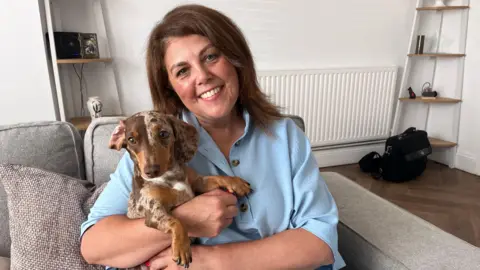 Glynda Cullen with her pet Dachshund, Lemon, sit on a sofa in her living room. Glynda has brown hair and wears a blue blouse. 