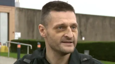 James Mee, who has short, brown hair and a stubbly beard is wearing a black jacket and T-shirt 