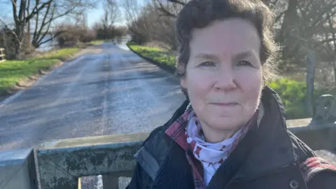 May Lowe has wavy black hair, she is wearing a dark coat with a pink and white scarf, behind her is a shut heavy metal barrier gate with a road beyond that, with water over the road in the far distance.