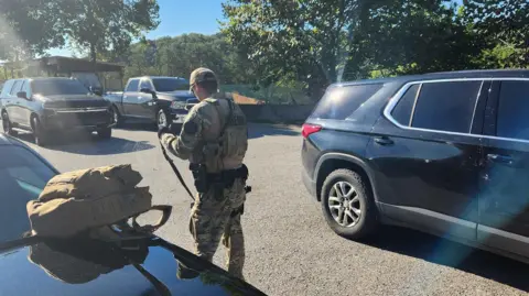 Laurel County Sheriff's Office. A man in tactical gear with a gun surrounded by cars on a highway.