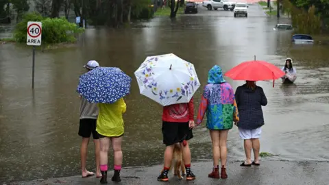 Residents in the Brisbane suburb of Newmarket carry umbrellas and stand on dry ground next to a flooded part of the road