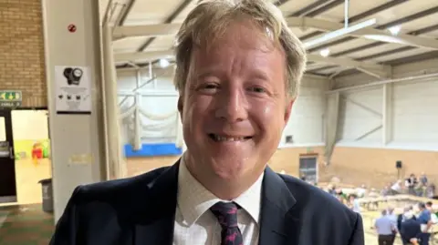 Paul Bristow at an election court in May 2025. He is smiling and wears a dark blue jacket with a white checked shirt and blue and red tie. 