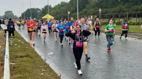 Submitted Miss Ahmed among dozens of runners taking part in Great North Run on a rainy day. She is wearing black running gear and is smiling and giving a thumbs up to the camera. 