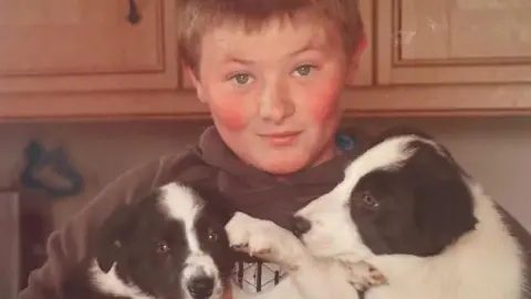 Jordan Burrow An old photo of Jordan. He is looking into the camera and holding two sheepdog puppies. His cheeks are flushed and he is wearing a brown hoodie.