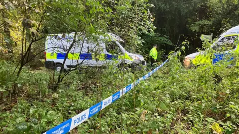 Two police vans facing each other in a wooded area, separated by police tape. There is a police office in front of one of the vans. 