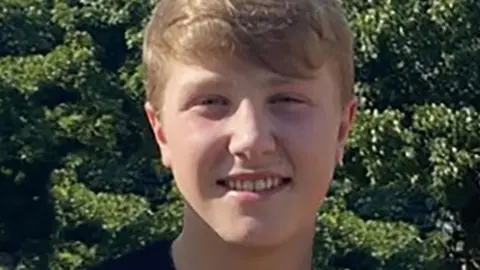 Matthew Daulby - a young man with blond hair wearing a black t-shirt, pictured smiling and standing in front of trees on a sunny day. 