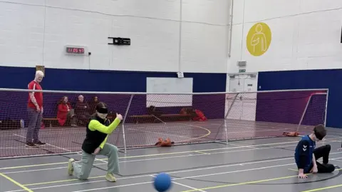 Leah Seabourne Goalball players are taking part in a taster session. They are wearing eyeshades. One player is on one knee after throwing the ball. Another player is sitting on the floor. Spectators are nearby. There is a net across the court.