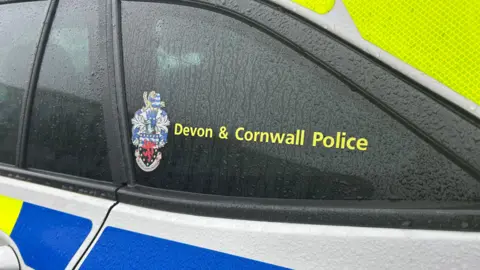 A close-up of the Devon and Cornwall Police logo on a police car. The vehicle has tinted windows and neon stickers. Rain droplets are running down the exterior of the car.
