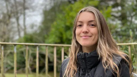 Laura is is seated outdoors and she is is wearing a black quilted vest over a long-sleeved top. Behind her is a railing and a garden area with green shrubs, trees, and grass, under an overcast sky.