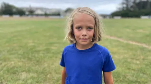 A boy with blond hair wearing a blue T shirt stands in a green field 