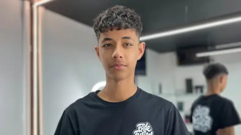 A boy is wearing a black t-shirt and has brown curly hair. He is standing in a barber shop with white walls and a black ceiling.