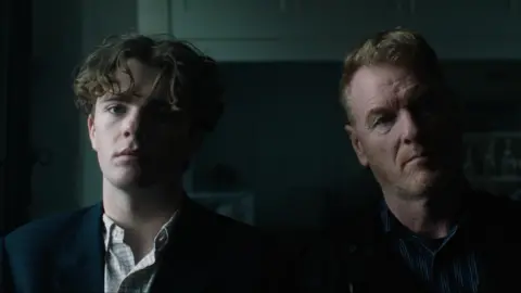 Max Morgan Two male actors, one younger than the other, stand next to each other in a dark room. Neither is smiling.