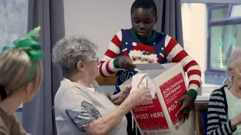 A boy handing out Christmas cards from a box to elderly residents