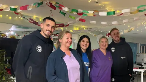 Captain Lewis Dunk, goalkeeper Jason Steele, and women's midfielder Dejana Stefanovic smile for the camera with two hospice staff wearing purple and pink scrubs under Christmas decorations from the ceiling and a Christmas tree to the side.