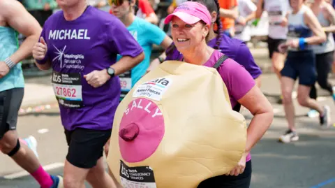 Sally Orange A woman wearing a giant breast costume which says "Sally" on it, smiles as she runs with others in the London Marathon.