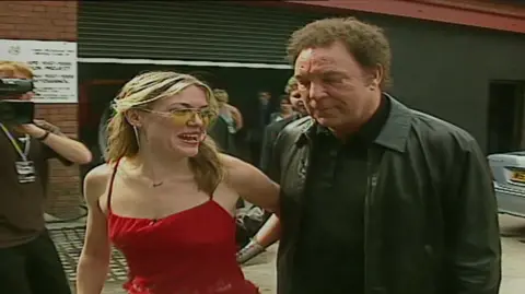 Cerys Matthews in a red dress and sunglasses with Tom Jones in a leather jacket. they arrived at the reopening of the Pop Factory in 2000.