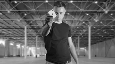 Greg Chipman A black and white photograph of a man with short slicked back hair wearing a black t-shirt. The man is in a huge warehouse building and is holding up in front of his body an ace of spades playing card.