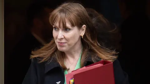 PA Media A close up photo of the Deputy Prime Minister Angela Rayner walking out of a building. She is wearing a black coat with a green shirt. She is also carrying a red folder with lots of paper inside. She is looking to her right. 