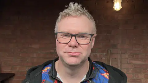 Sam Read Close up of Rich Hall standing in front of a brick wall. He is wearing a hooded top and has spiky grey hair.