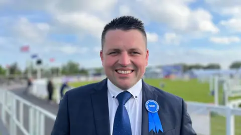 Richard Beattie. He has shirt dark hair, wearing a navy blazer, white shirt, blue tie and blue rosette pin.