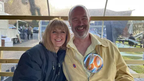Alyson Noakes Alyson and Stephen sit together in a small wooden hut. Alyson is wearing a dark grey coat while Stephen, in a yellow shirt and yellow jacket with 60th birthday badges on, smiles. He has a partially bald head and a short grey beard. 