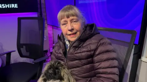 Woman sat in radio studio with dog sat on her lap smiling to camera.