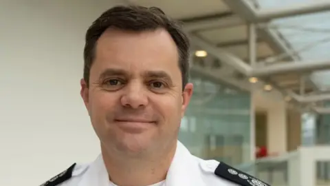 Suffolk County Council A man with greying brown hair and brown eyes looks directly into the camera with a half smile on his face. His white collared shirt has black fire service epaulettes.