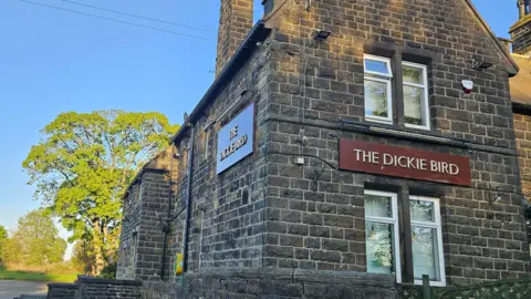 Aisha Iqbal/BBC A pub building, built from heritage stones. It has maroon signs with gold writing identifying it as 'The Dickie Bird'. 