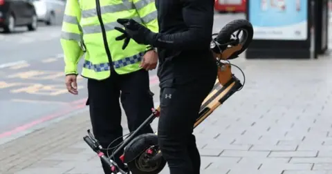 PA Media A generic image features an unidentified police officer, dressed in a hi-vis jacket talking to an e-scooter user, who is carrying his machine and is dressed in black. They are standing on a wide pavement, with a road and a bus shelter in the background.