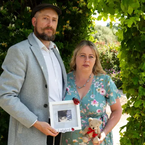 BBC/Chris Waring Carl Everson and Carly Wesson stand in a garden holding an ultrasound picture, in a white frame, and a teddy wearing a red ribbon.