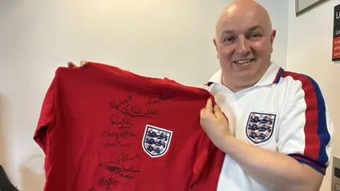 A bald man wearing an old England football shirt holds up a red 1966 World Cup winning shirt with signatures on the front