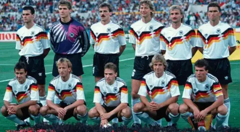 Getty Images The 1990 German football team stand in a two row line-up in the tournament. One row is squatting with the other stood behind. Their tops are white with yellow, black and red zig zag stripes on with black shorts.