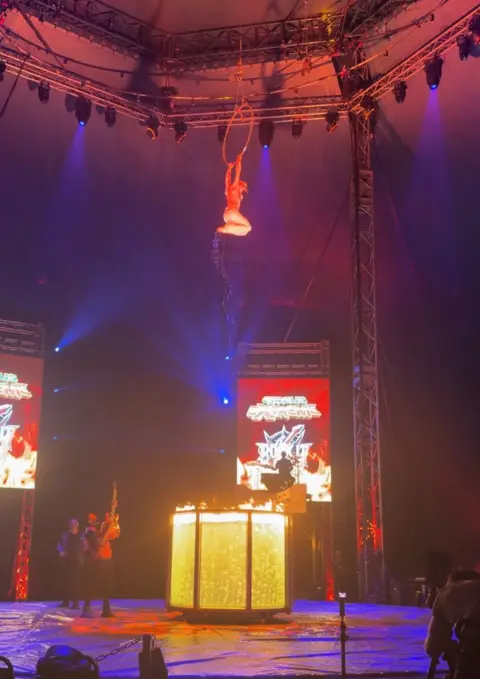 Laura about to fall feet first from high up on the big top tent, in to a see through container of water with flames on the surface of the water.