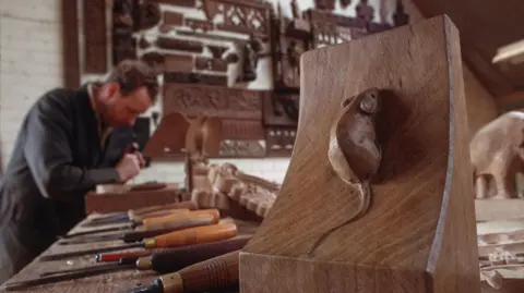 Getty Images A detailed wooden carving of a small mouse on a slanted block in the foreground, surrounded by woodworking tools on a bench. The background shows a workshop filled with intricate wooden carvings mounted on the wall and other wooden pieces on the work surface.
