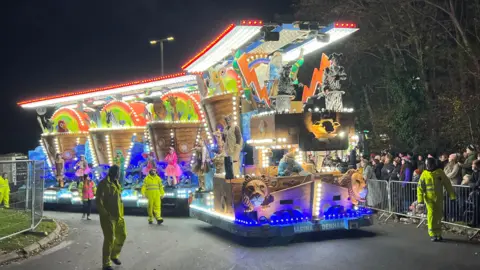 An illuminated colourful carnival cart. There is a crowd to the side of it and people walking alongside it wearing high-vis jackets. The float has illustrations of animals on it.