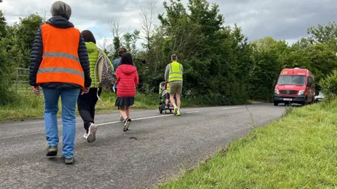 George Carden/BBC Adults, one who is wearing a high vis vest, and two children wearing high vis crossing Chalkers Lane with traffic letting them get across. One person is pushing a pram 