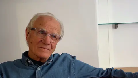 Head and shoulders shot of John Henwood sitting wearing a denim shirt and glasses in front of a white wall