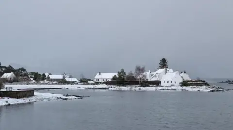 BBC Weather Watchers/happydays Snow covered houses in a little coastal town surrounded by water on a grey day.