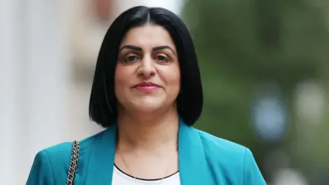 EPA Shabana Mahmood is wearing a teal jacket and white shirt with a gold chain and we can see a gold bag chain. She has shoulder-length, straight black hair and is smiling to camera while on a street. The background is out of focus.