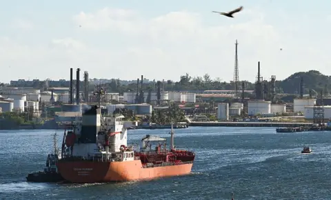 A container ship sails along choppy waters near a sprawling industrial facility close to the shore. It is daytime and a bird flies over the water.