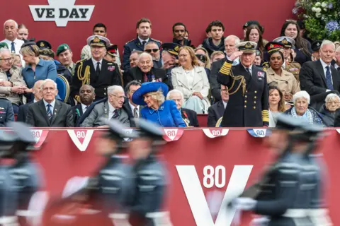 Reuters The same moment, the King saluting, is shown from the other side - the King can be seen front-on as he salutes wearing formal attire surrounded by other members of the crowd sitting while military personnell march in the foreground.